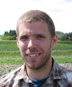 Ian Epp smiles while having his portrait taken. He is standing outside with a field behind him and is wearing a brown checkered shirt. 