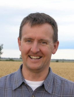 Tom Wolf smiles as he has his photo taken. He stands in front of a vast field and is wearing a blue checkered shirt. 