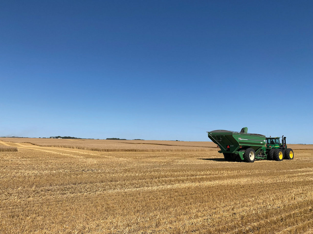 A green tractor with a grain cart attachment in the middle of a vast, harvested field under a clear blue sky.
